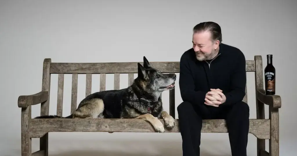 Ricky Gervais sitting on a bench looking at a dog smiling with a bottle of Dutch Barn Vodka next to him